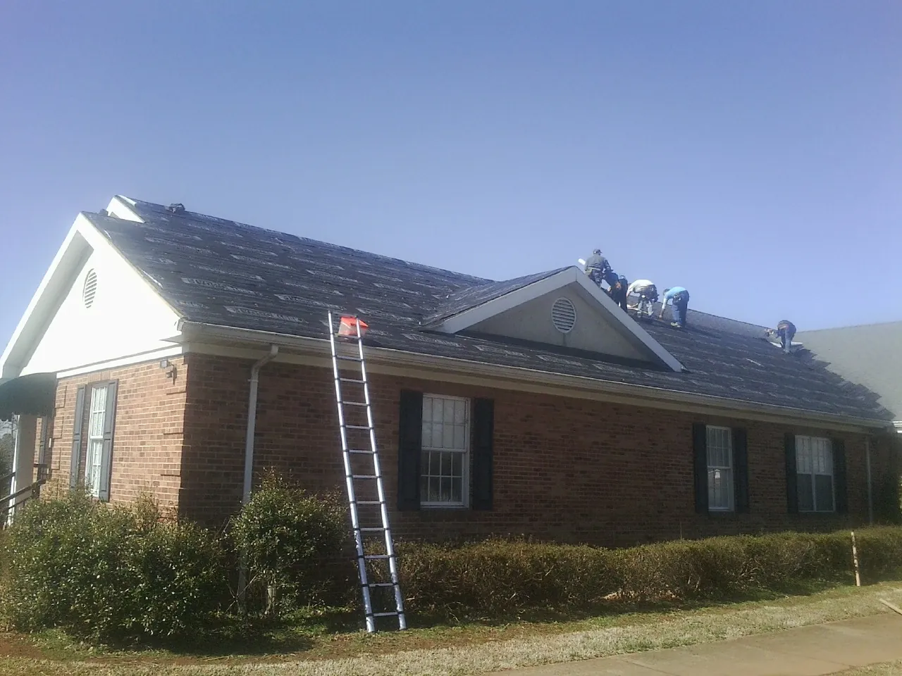 Crew tearing shingles off a brick ranch roof with a ladder set — Metal Roof Repair work by our crew in Mount Sinai
