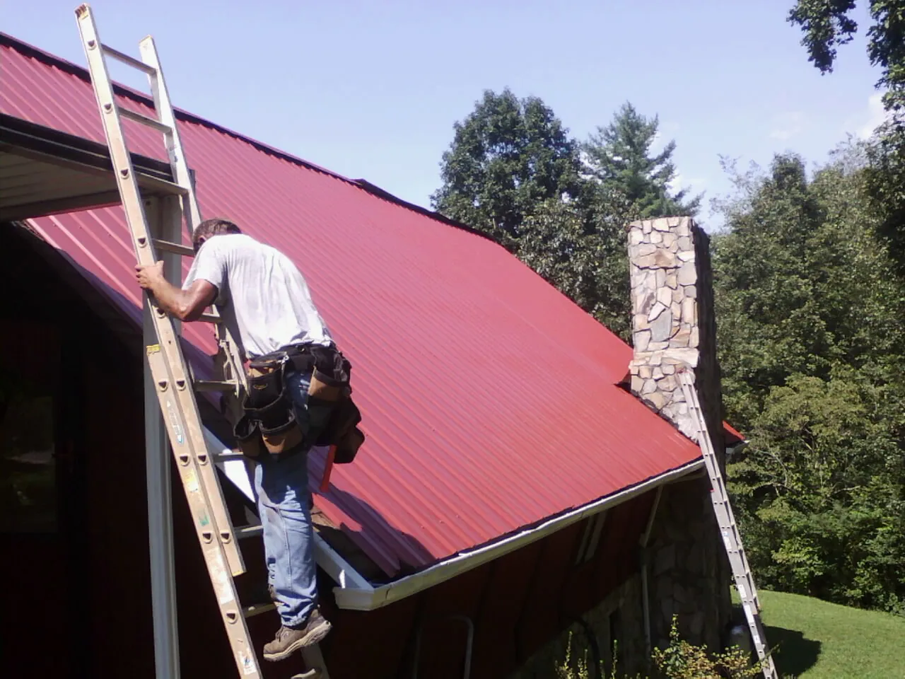 Roofer on a ladder beside a red metal roof and stone chimney — Soffit & Fascia Repair by Roofing Brothers in Mount Sinai